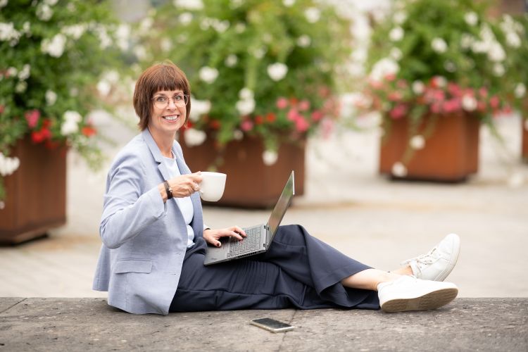 Monika Biewer Porträtfoto freundlich lächelnd nach vorne schauend. Sie sitzt auf einer Bank, hält eine Tasse Kaffee in der Hand und ihr Laptop liegt aufgeklappt auf ihrem Schoß